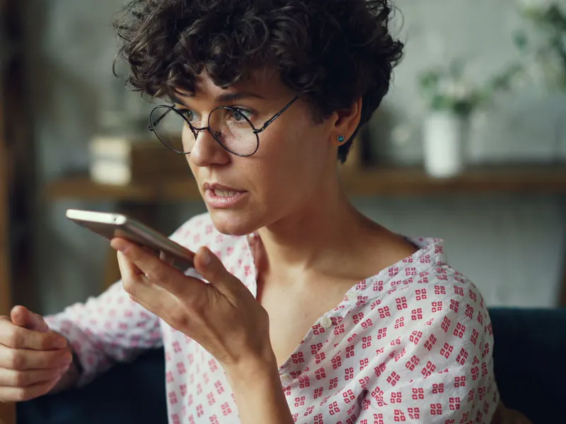 Woman with curly hair and round glasses speaking into a smartphone held near her mouth at home