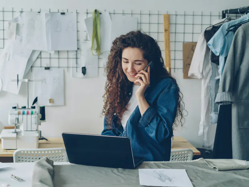 Small business owner in her clothing studio answering a phone call while working on a laptop