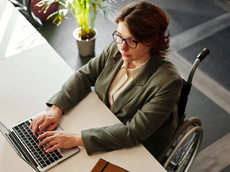 Woman in glasses and an olive blazer typing on a laptop at a white desk while seated in a wheelchair, seen from above