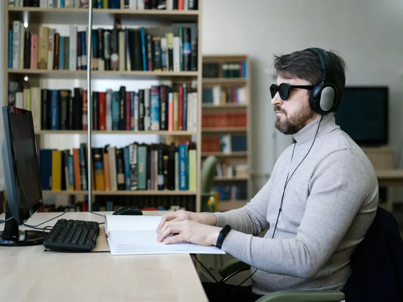 Man wearing dark glasses and over-ear headphones reading a braille sheet with one hand at a desk with a keyboard and monitor