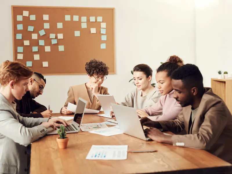 Six coworkers working on laptops and papers around a wooden table, with a corkboard full of sticky notes on the wall behind