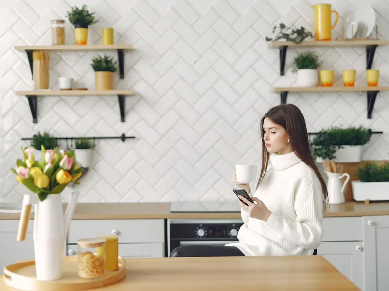 Young woman in a white sweater holding a coffee mug and looking up something on her phone at a bright kitchen counter