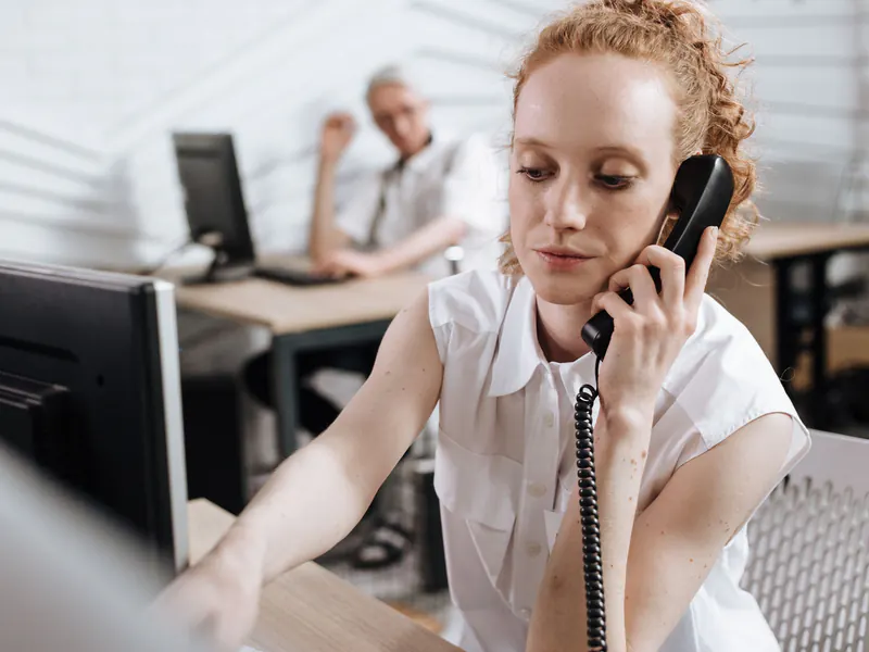 Woman with curly red hair on a corded desk phone at her computer, a colleague working at another desk behind her in an open office