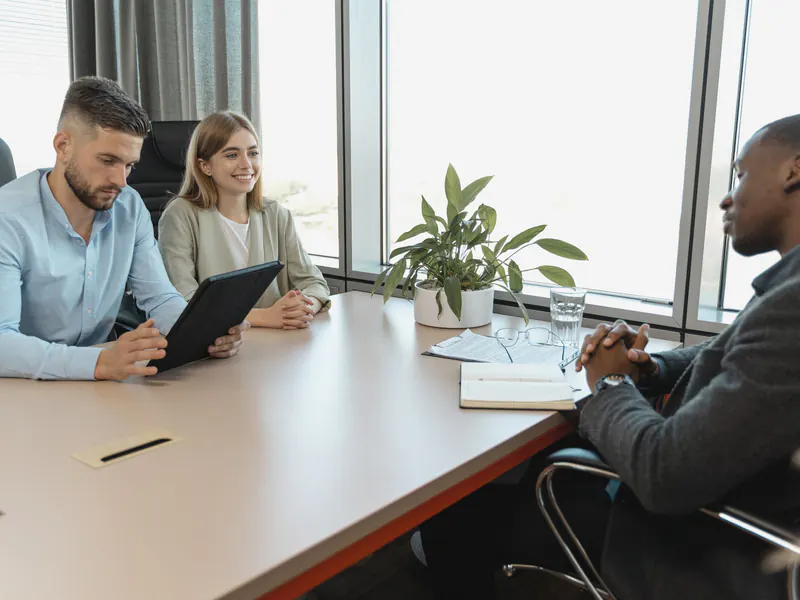 Two interviewers, a man with a tablet and a smiling woman, face a candidate in a gray blazer across a meeting table