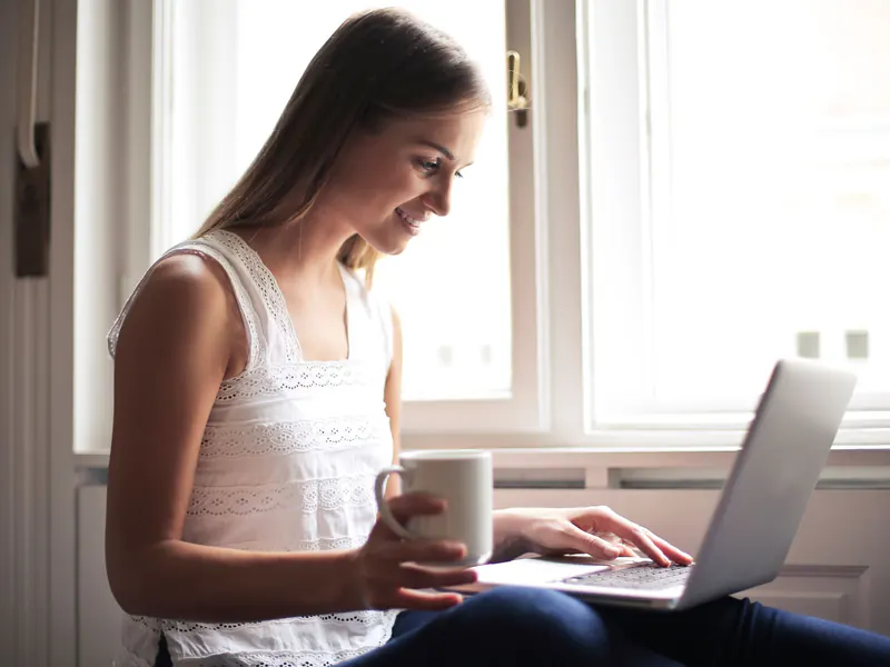 A smiling woman holds a coffee mug and types on a laptop on her lap beside a sunlit window at home.