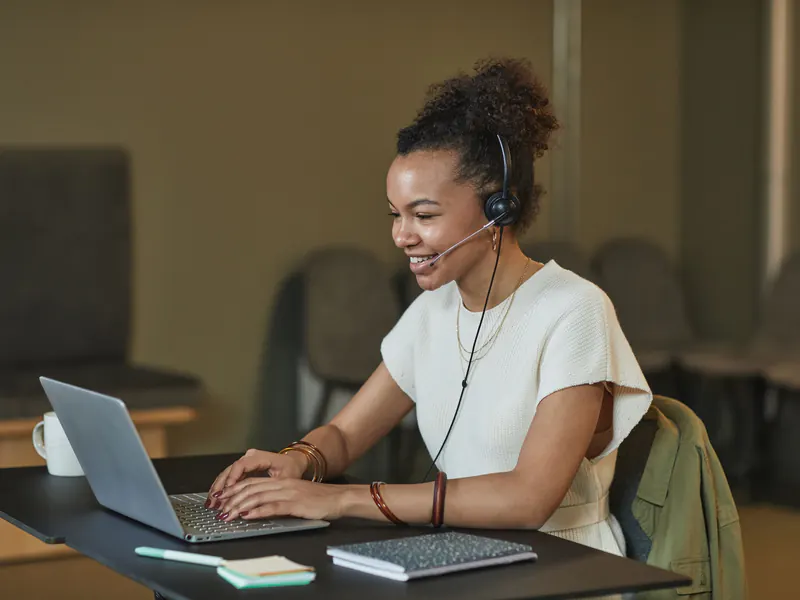 Smiling woman wearing a headset with a microphone, typing on a laptop at an office desk with a notebook and sticky notes