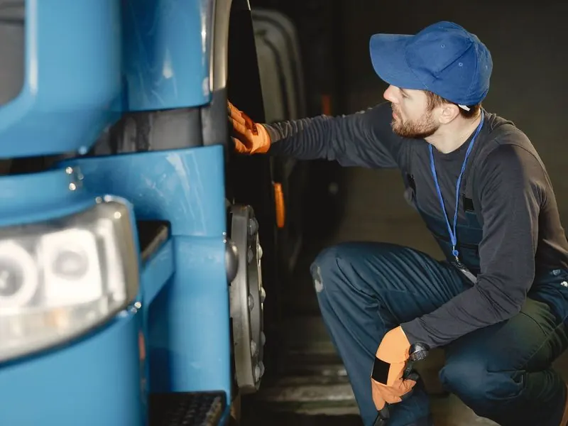 Worker in a blue cap and work gloves crouching to inspect the front wheel of a blue truck