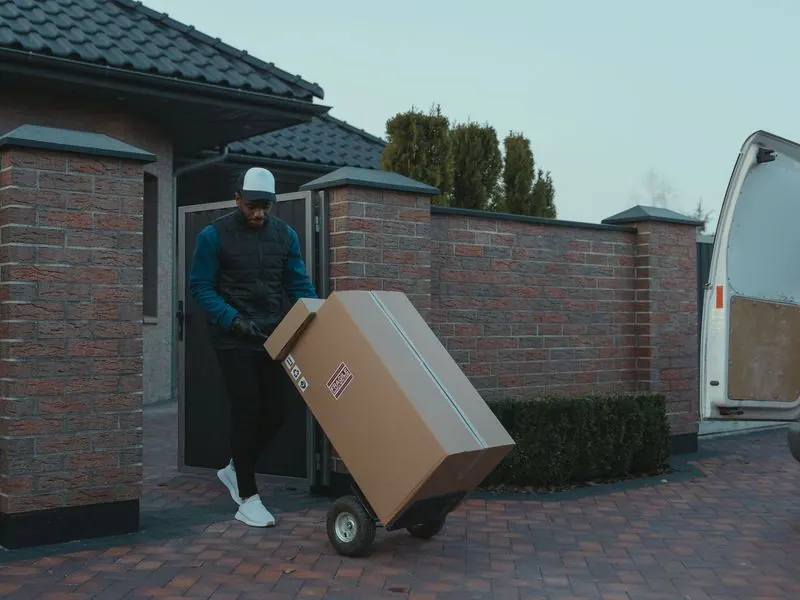 Delivery driver wheeling a large boxed parcel on a hand truck out of a gated driveway toward an open van