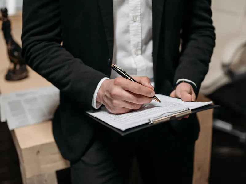 Close-up of a man in a dark suit signing a printed agreement on a black clipboard with a fountain pen in an office