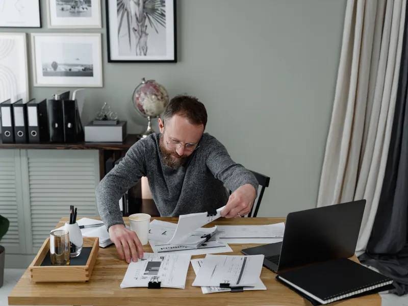Bearded man in glasses and a gray sweater reviewing printed documents spread across his desk beside an open laptop