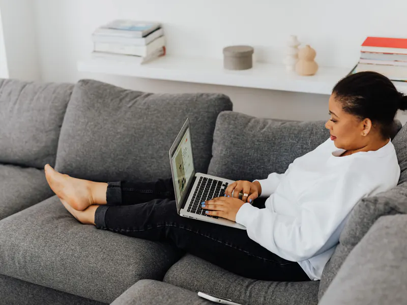 Woman in a white sweatshirt relaxing barefoot on a gray sofa, typing on a laptop in a bright living room