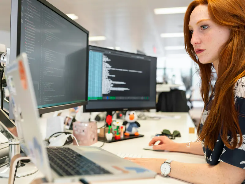A developer with red hair works at a desk with two monitors showing code and a laptop in a bright office.