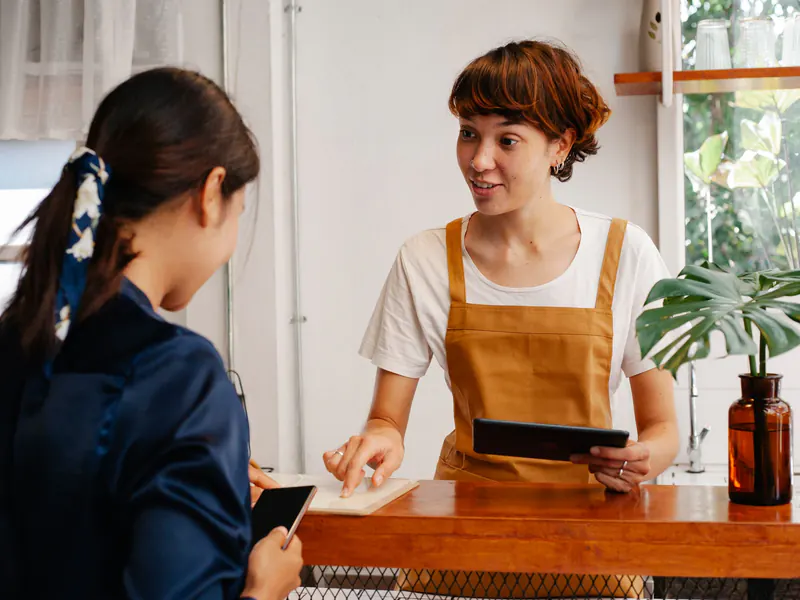 A cafe worker in an orange apron holds a tablet and talks with a customer across a wooden counter.