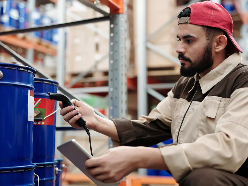 Warehouse worker in a red cap crouching between shelves, scanning blue barrels with a handheld scanner