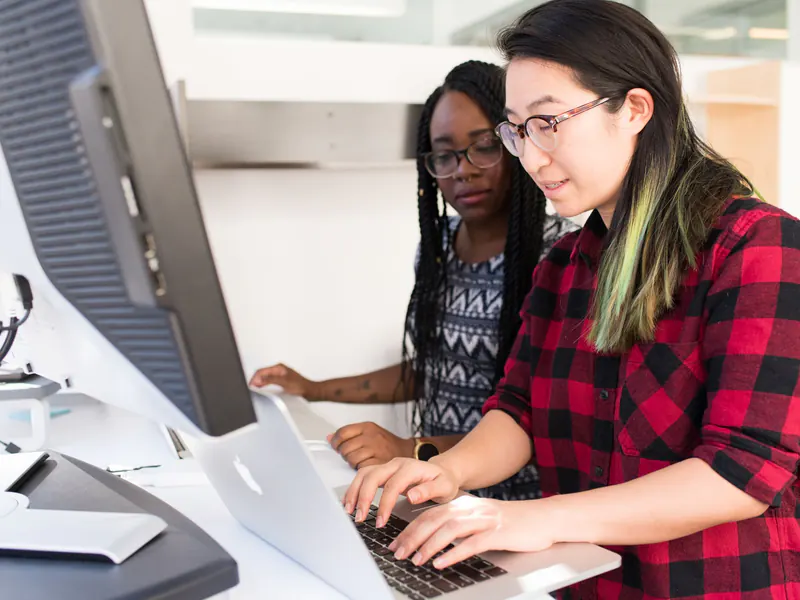 Two women developers working together at one desk, one typing on a laptop while the other leans in to follow along