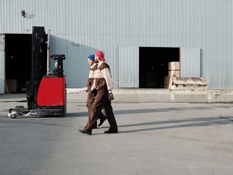 Two warehouse workers in uniforms walking past open loading-dock doors beside a red forklift on a sunny yard