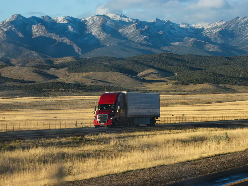 A red semi truck with a white trailer drives along a highway through grassland below snow-capped mountains