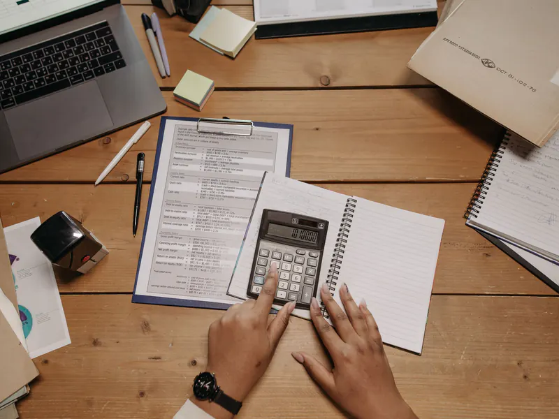 Top view of hands using a desk calculator over a notebook, beside a clipboard of figures and an open laptop