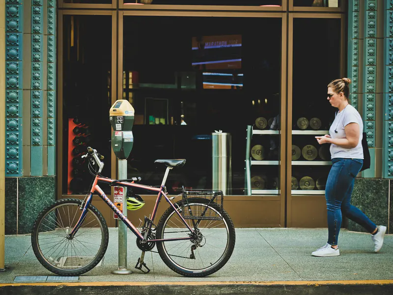 Woman in a gray T-shirt walking along a downtown sidewalk while checking her phone, passing a shop window