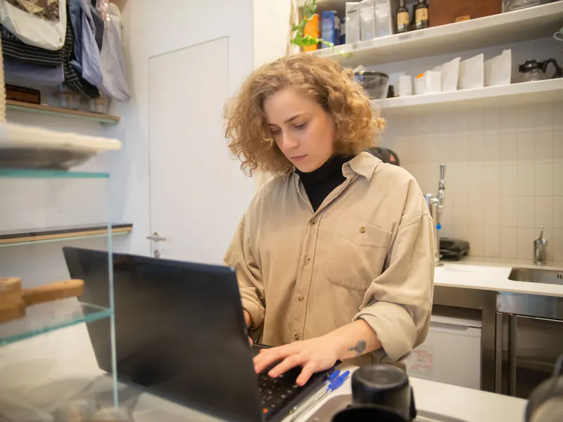 Curly-haired woman in a beige shirt typing on a laptop at the counter of her small shop, with shelves of supplies behind her