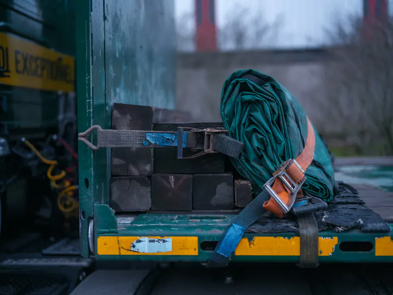 Close-up of orange and grey ratchet straps cinching a rolled tarp and wooden dunnage blocks on a worn flatbed trailer deck