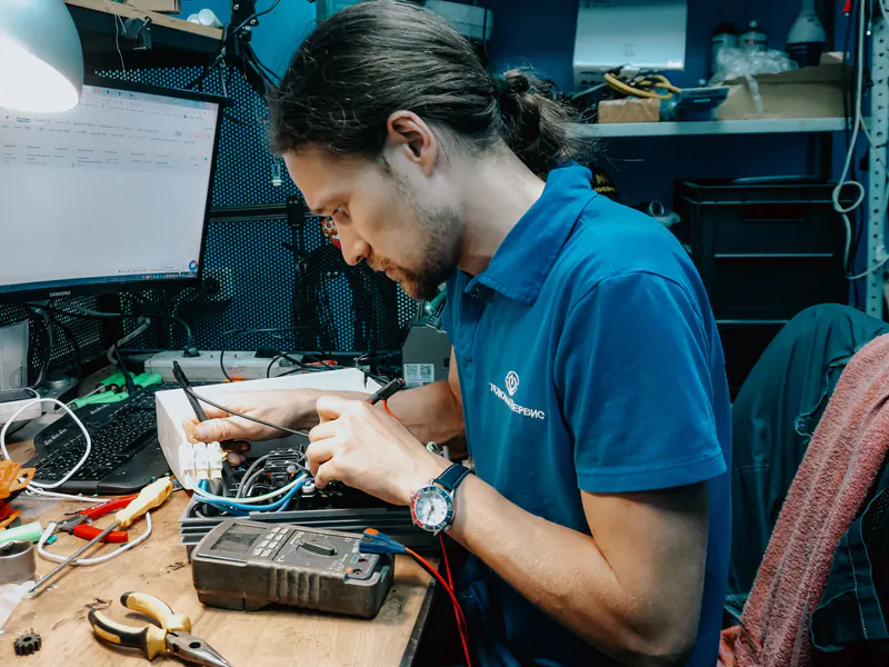 Technician in a blue polo shirt repairing electronics at a cluttered workbench with a multimeter, pliers and wiring