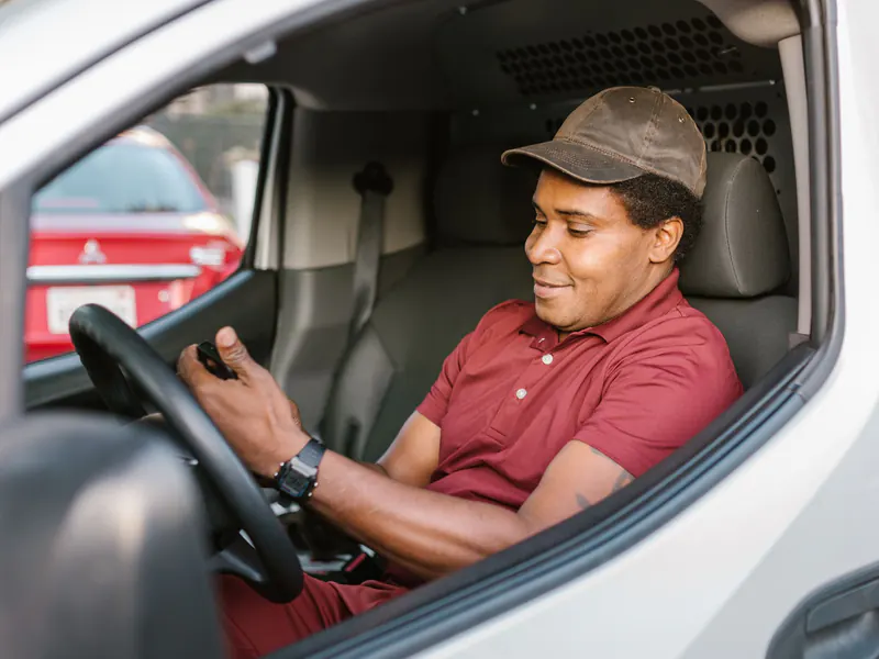 Smiling driver in a red polo shirt and brown cap seated behind the wheel of a white van, looking down at a phone in his hand