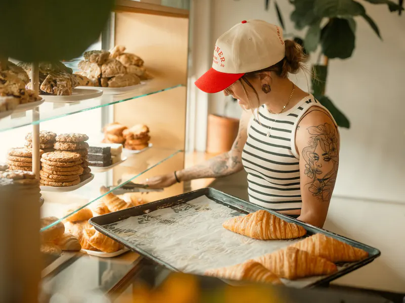 Baker in a cap and striped tank top setting a tray of fresh croissants into a glass display case filled with cookies