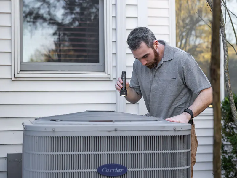 Bearded technician in a gray polo shining a flashlight into an outdoor air-conditioning condenser beside a house