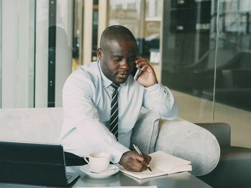A man in a shirt and tie talks on the phone while writing in an open notebook beside his laptop.