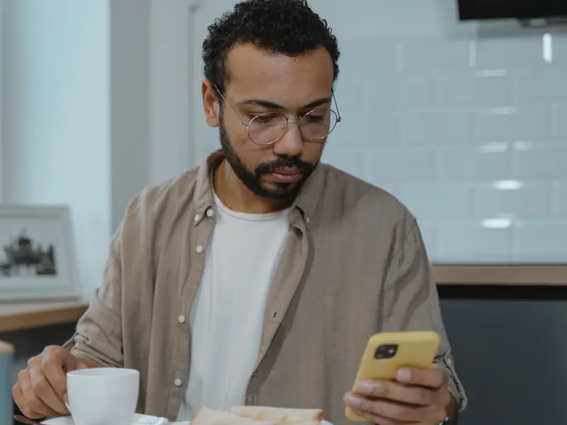 Bearded man in glasses reading something on his smartphone at a kitchen table at home, holding a coffee cup