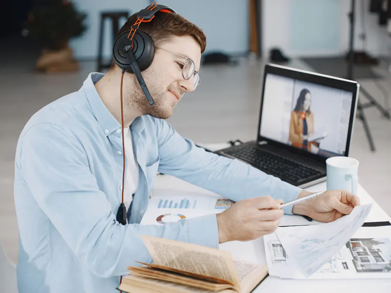 Man in a headset marking up printed papers at his desk while a woman appears on a video call on his laptop