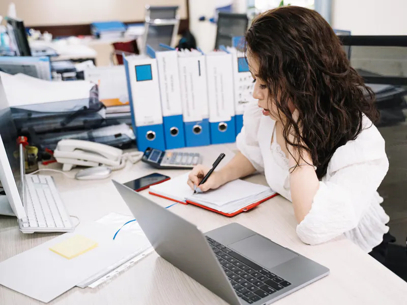 Woman writing in a notebook at an office desk beside a laptop, with binders, a calculator and a desk phone around her