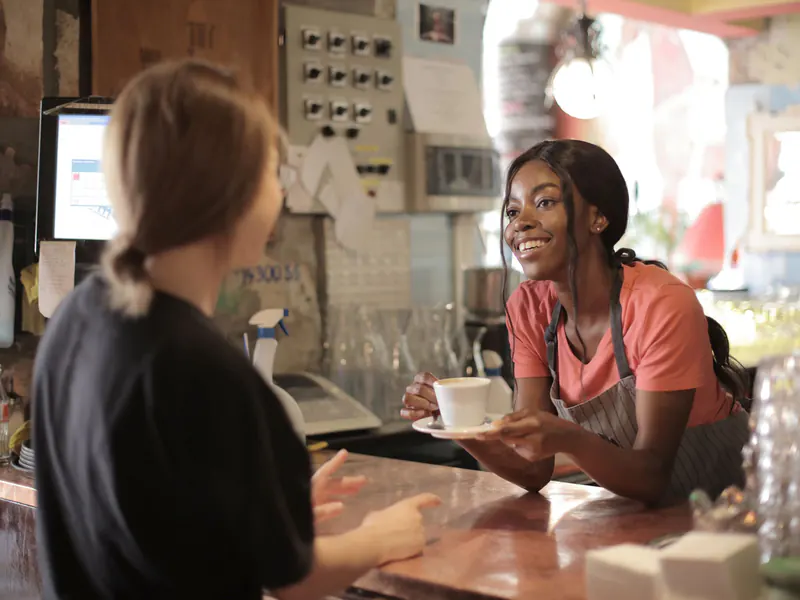 Smiling barista in an apron handing a cup of coffee across the counter to a customer in a busy, cozy cafe