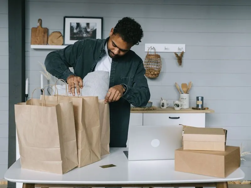 Man in a denim shirt packing an order into kraft paper bags beside a laptop and shipping boxes on a white table