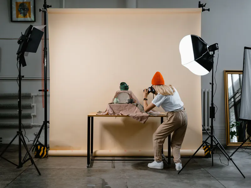 Photographer in an orange beanie shooting a product on a small table against a paper backdrop, lit by a softbox