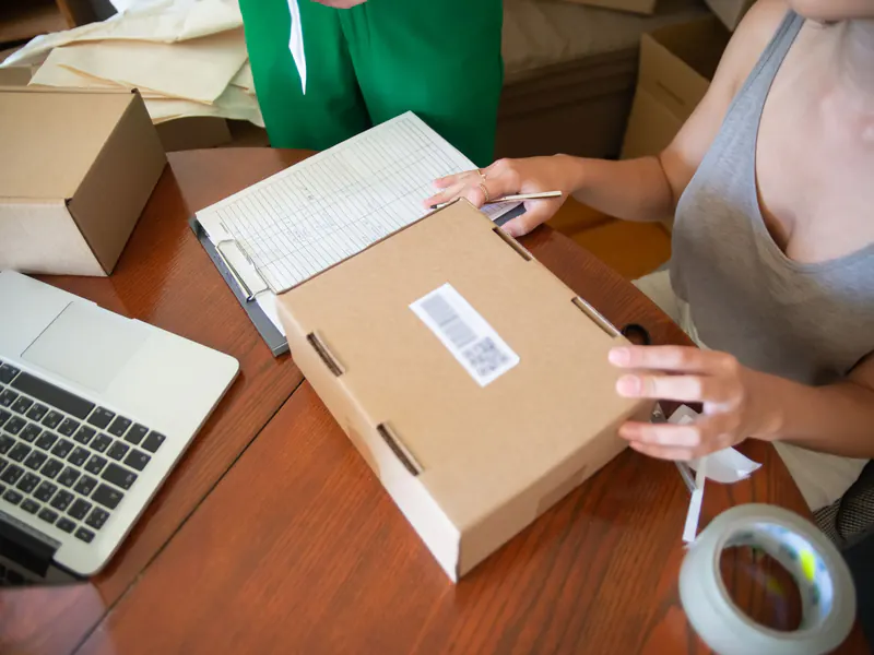 Woman sealing a shipping box with a printed label beside a roll of tape, an order sheet and an open laptop on a wooden table