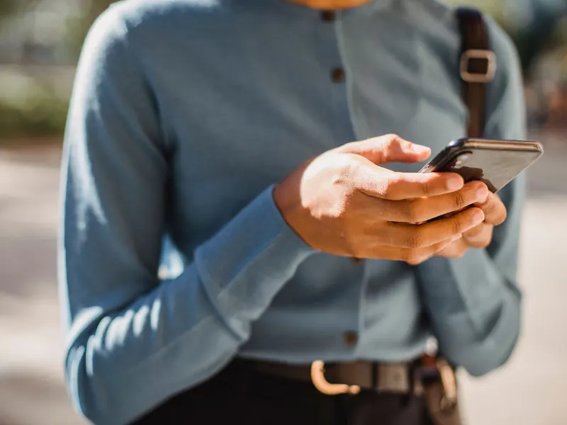 Close-up of a man in a blue button-down shirt with a bag strap holding a smartphone outdoors, screen turned away from view