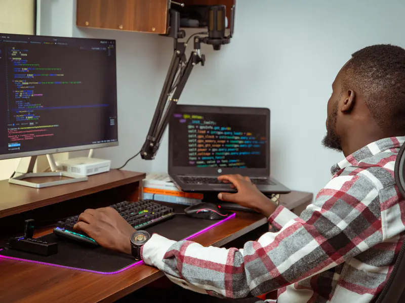 Developer in a plaid shirt typing at a desk with code on a large monitor and a second laptop beside a mechanical keyboard