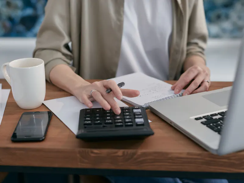 Hands working a desk calculator beside a notepad, phone, coffee mug and open laptop on a wooden table while planning a budget