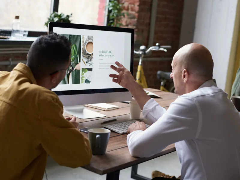 Two men at a wooden desk reviewing a website layout on a large desktop monitor, one gesturing at the screen while they talk