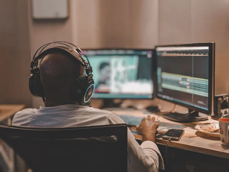 Man in headphones seen from behind at a desk, editing video across two monitors with a soft blurred timeline, warm room light