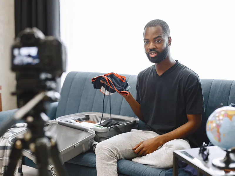 Man on a living-room sofa talking to a camera on a tripod while holding up clothes from an open suitcase