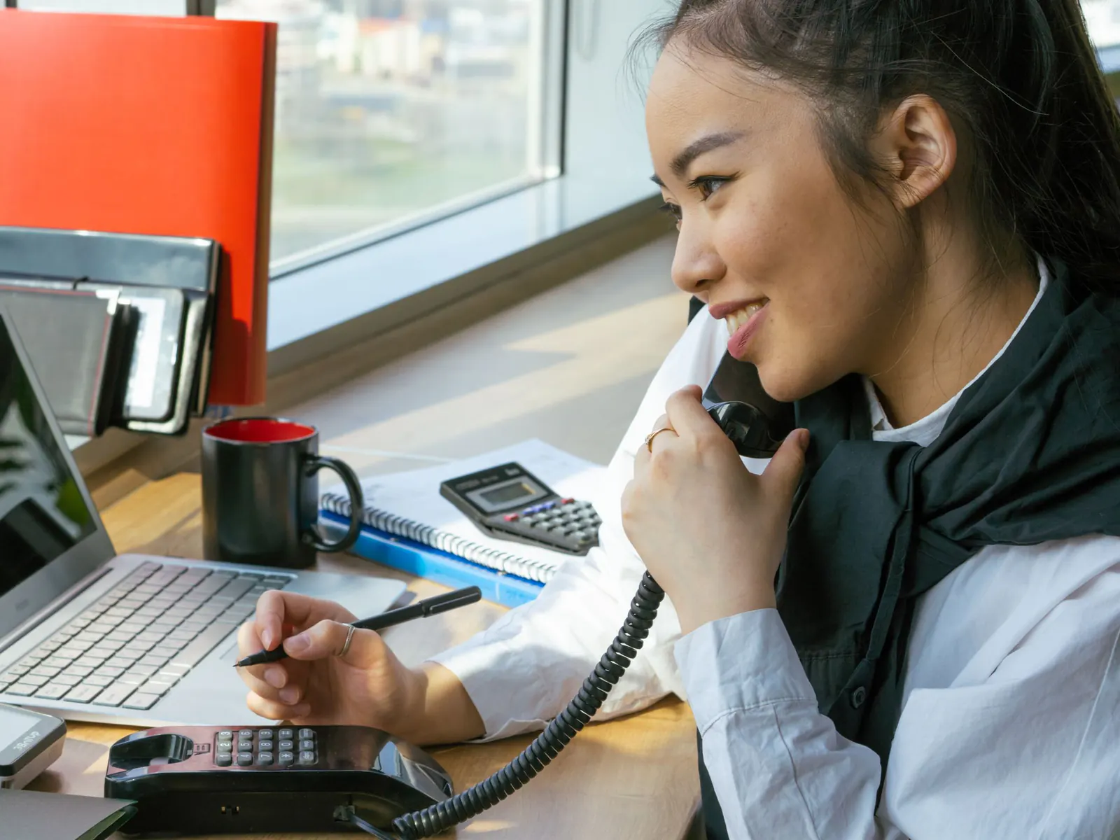 Smiling woman on a corded desk phone taking notes at a sunny office desk with a laptop and calculator
