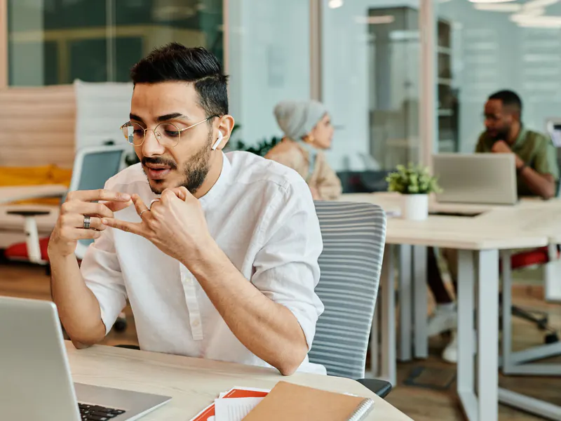 Man wearing wireless earbuds talking and gesturing during a call at his laptop in an open-plan office