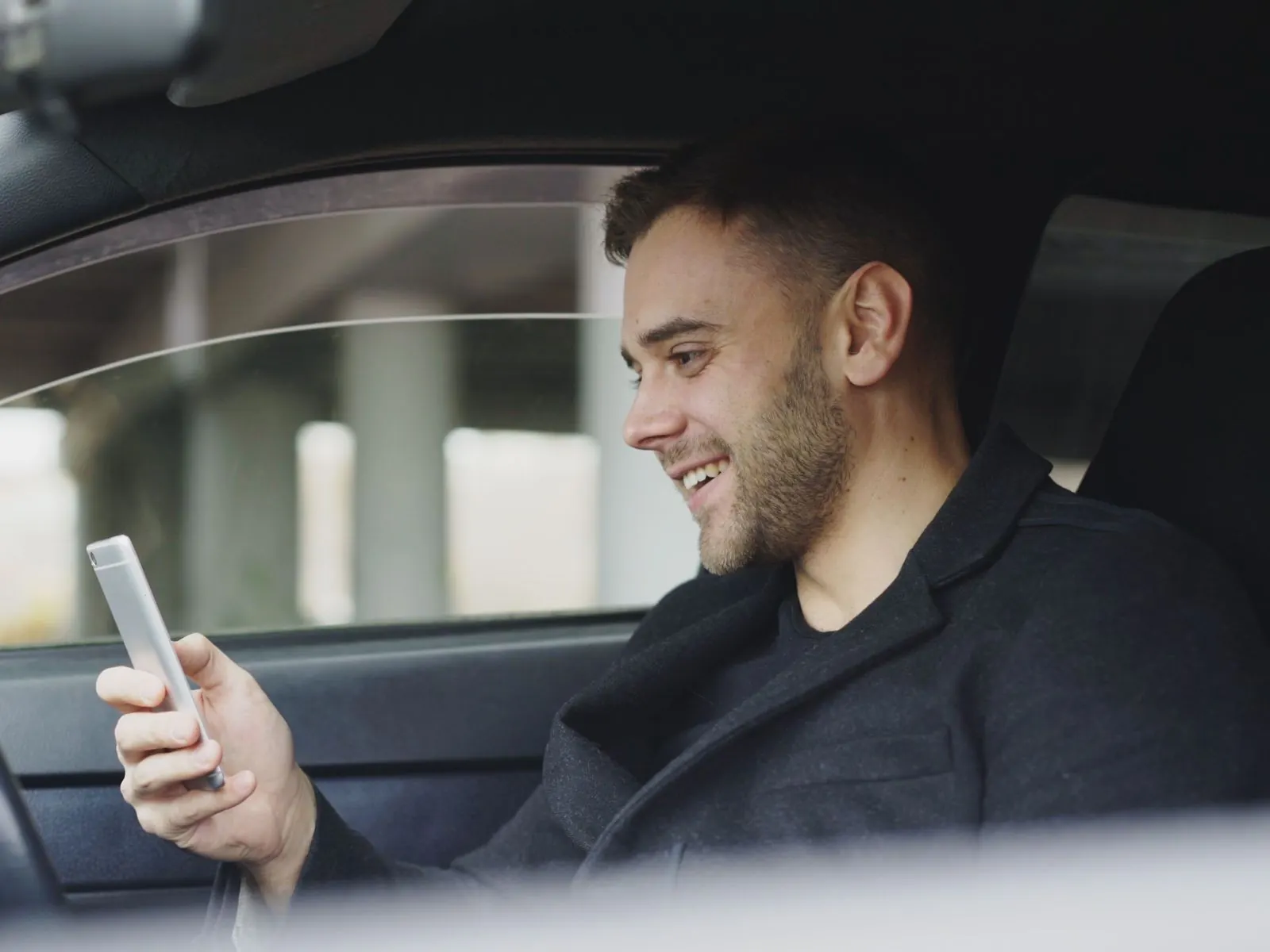 Young bearded man in a dark jacket sits in the driver's seat of a car, smiling as he looks at the smartphone in his hand