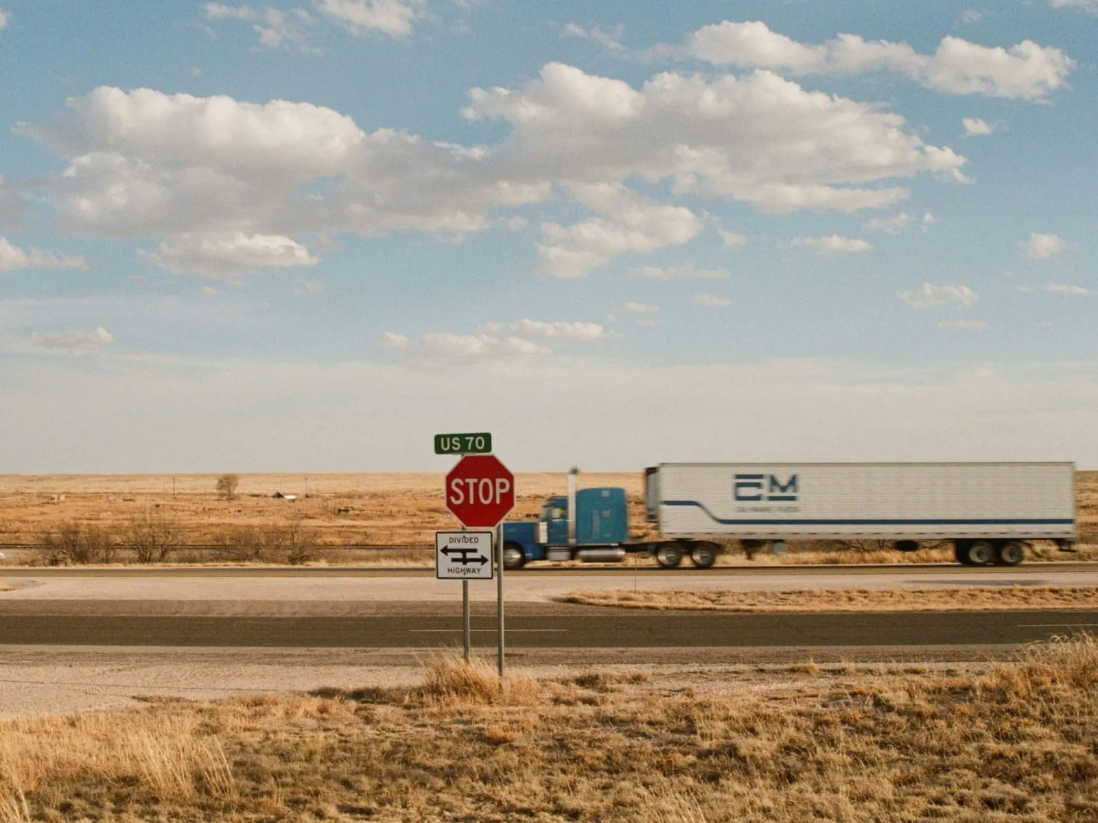 Blue semi-truck pulling a white trailer across flat, dry grassland behind a STOP sign and a US 70 route marker under a wide, cloudy sky
