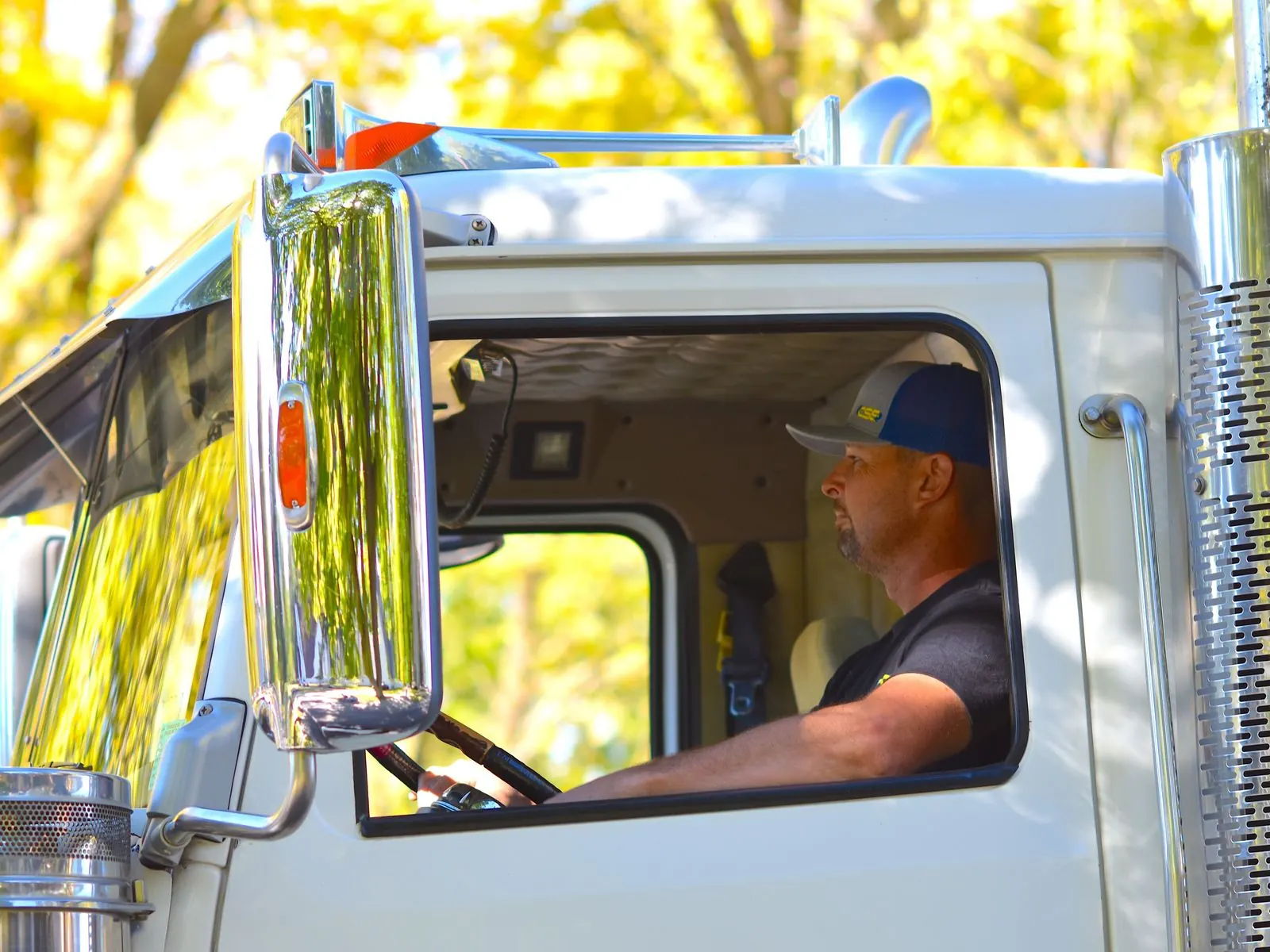 Truck driver in a baseball cap sits at the wheel of a white semi-truck, seen through the side window next to a large chrome mirror and exhaust stack