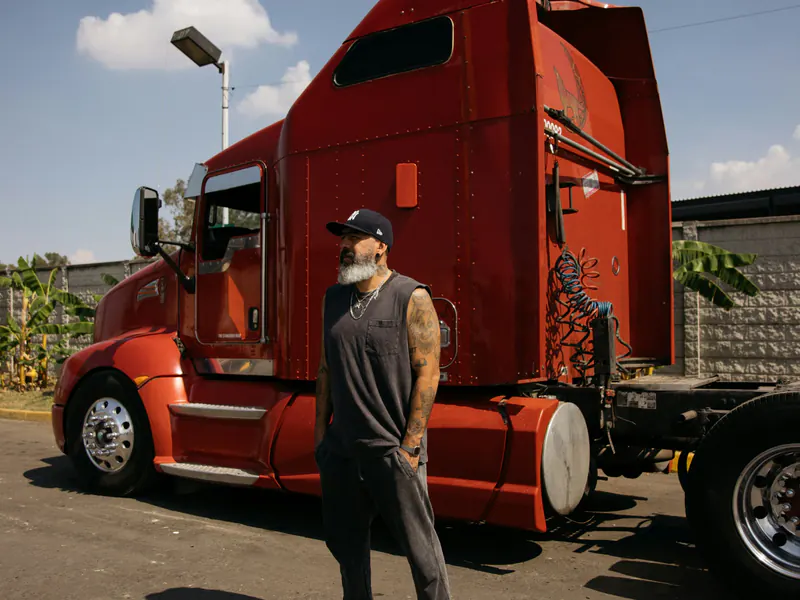 Bearded man in a cap and sleeveless shirt standing beside a red long-nose semi tractor on a sunny paved lot