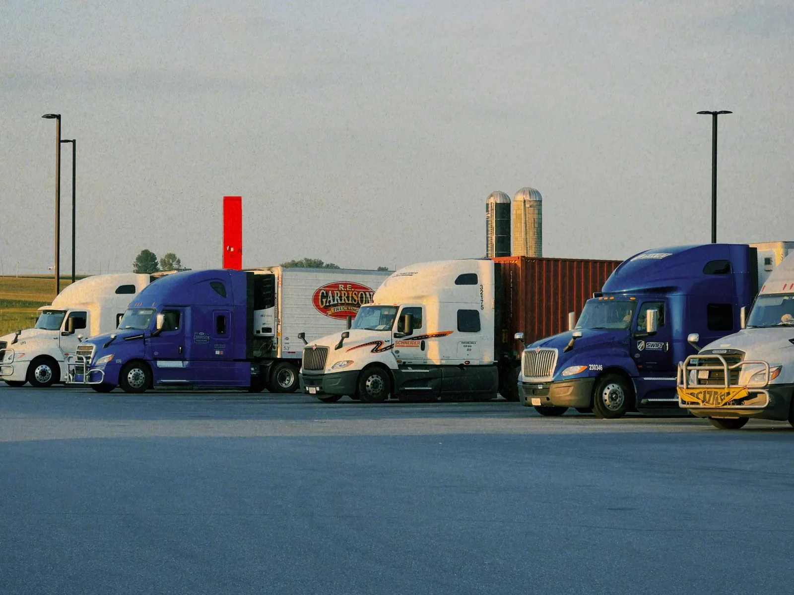 Row of semi-trucks parked side by side in a wide paved lot under a pale sky, with farm fields, grain silos and tall light poles behind